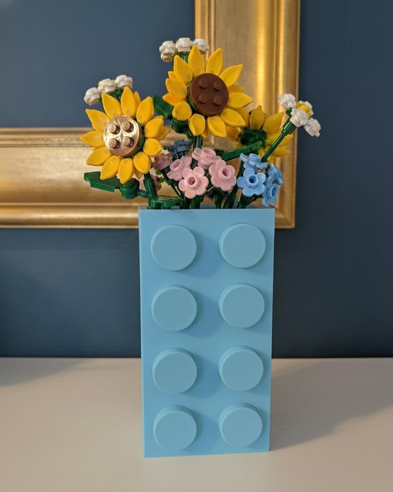 May include: A light blue vase made of interlocking blocks holds a bouquet of colorful flowers. The flowers include yellow sunflowers, pink and blue blossoms, and white accents. The vase is on a white surface.