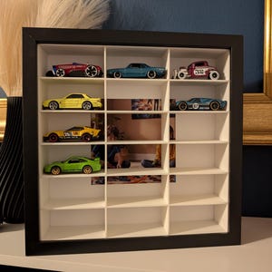 May include: A black framed display case with multiple compartments, showcasing a collection of toy cars. The cars are various colors, including red, blue, yellow, and green. The case is on a white surface, with a blue wall in the background.