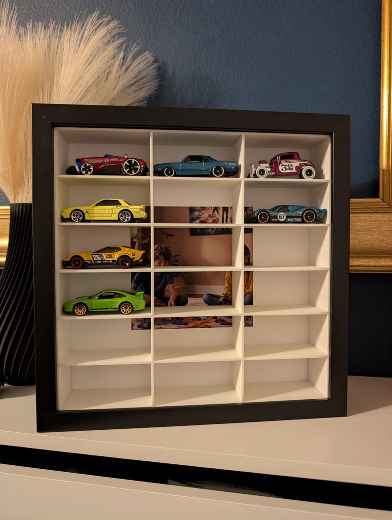 May include: A black framed display case with multiple compartments, showcasing a collection of toy cars. The cars are various colors, including red, blue, yellow, and green. The case is on a white surface, with a blue wall in the background.