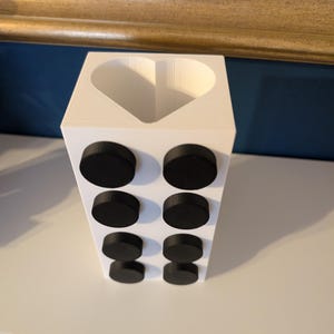 May include: A white rectangular container with a heart-shaped opening at the top. Eight black, circular magnets are attached to the front of the container. The container is sitting on a white surface.