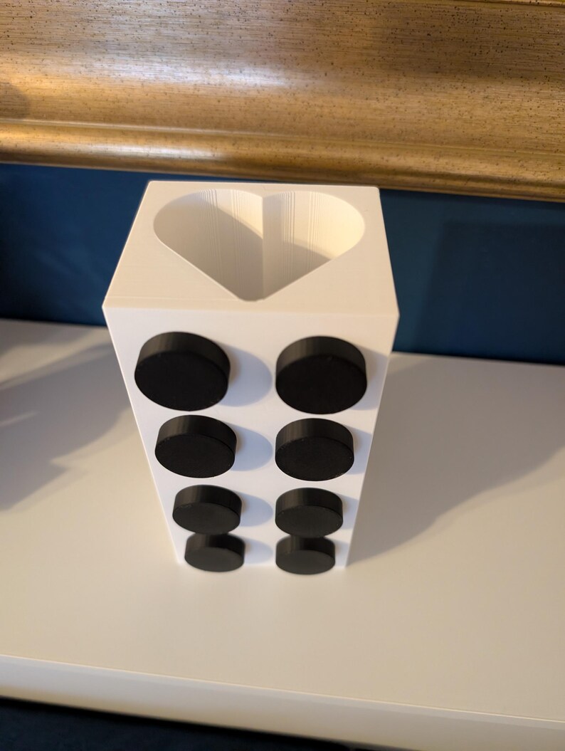 May include: A white rectangular container with a heart-shaped opening at the top. Eight black, circular magnets are attached to the front of the container. The container is sitting on a white surface.
