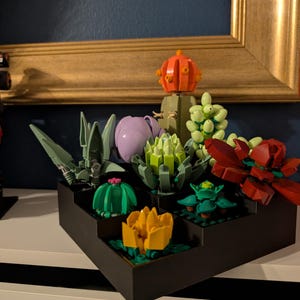 May include: A black tiered planter filled with colourful succulent plants made from toy building blocks. The succulents are various colours, including green, red, yellow, and purple. The background includes a gold-framed picture and a dark blue wall.