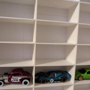 May include: A white display shelf with multiple compartments, each holding a toy car. The cars are various colors, including yellow, purple, blue, and green. The cars have different designs and numbers on them.