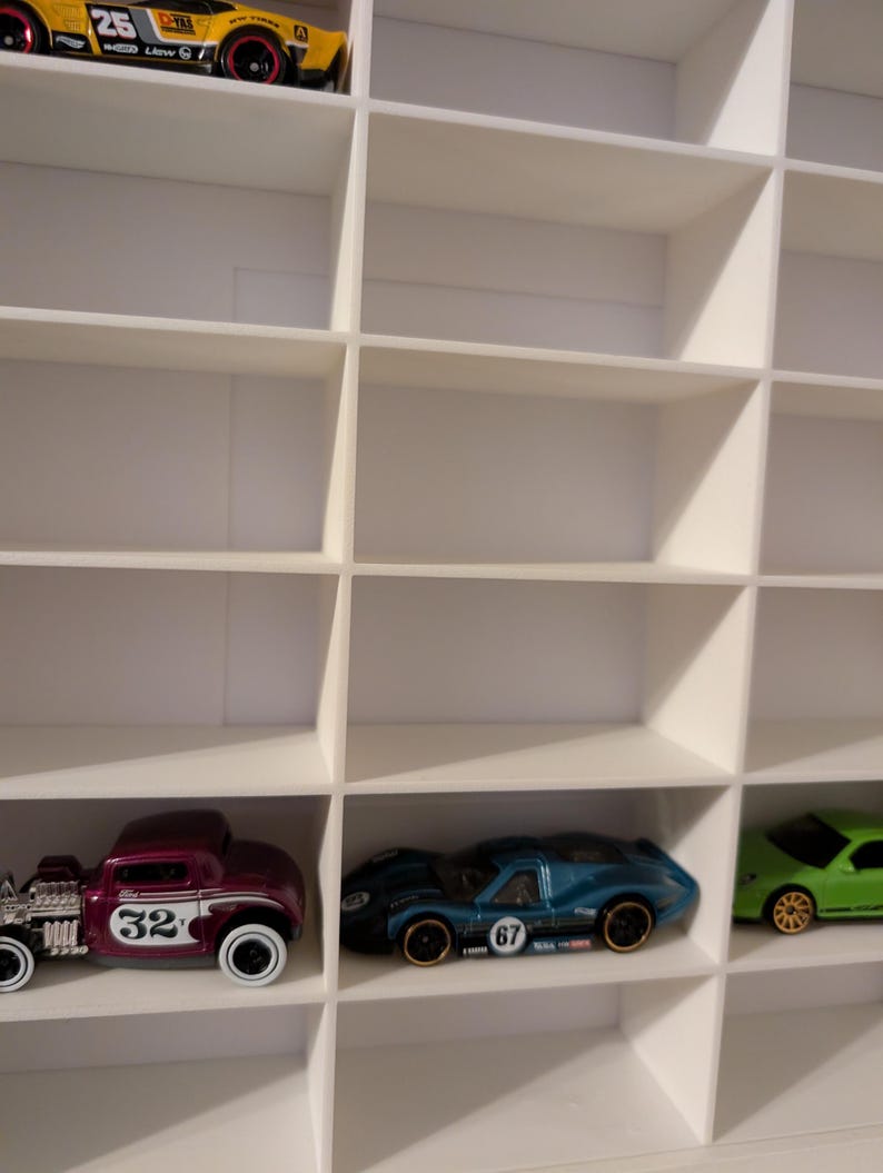 May include: A white display shelf with multiple compartments, each holding a toy car. The cars are various colors, including yellow, purple, blue, and green. The cars have different designs and numbers on them.