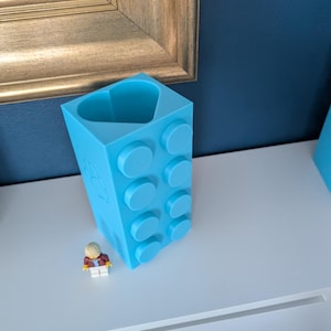 May include: A light blue, 3D-printed block-shaped container with circular studs, resembling a large building brick. A small Lego figure stands nearby. The container has a heart-shaped opening at the top.