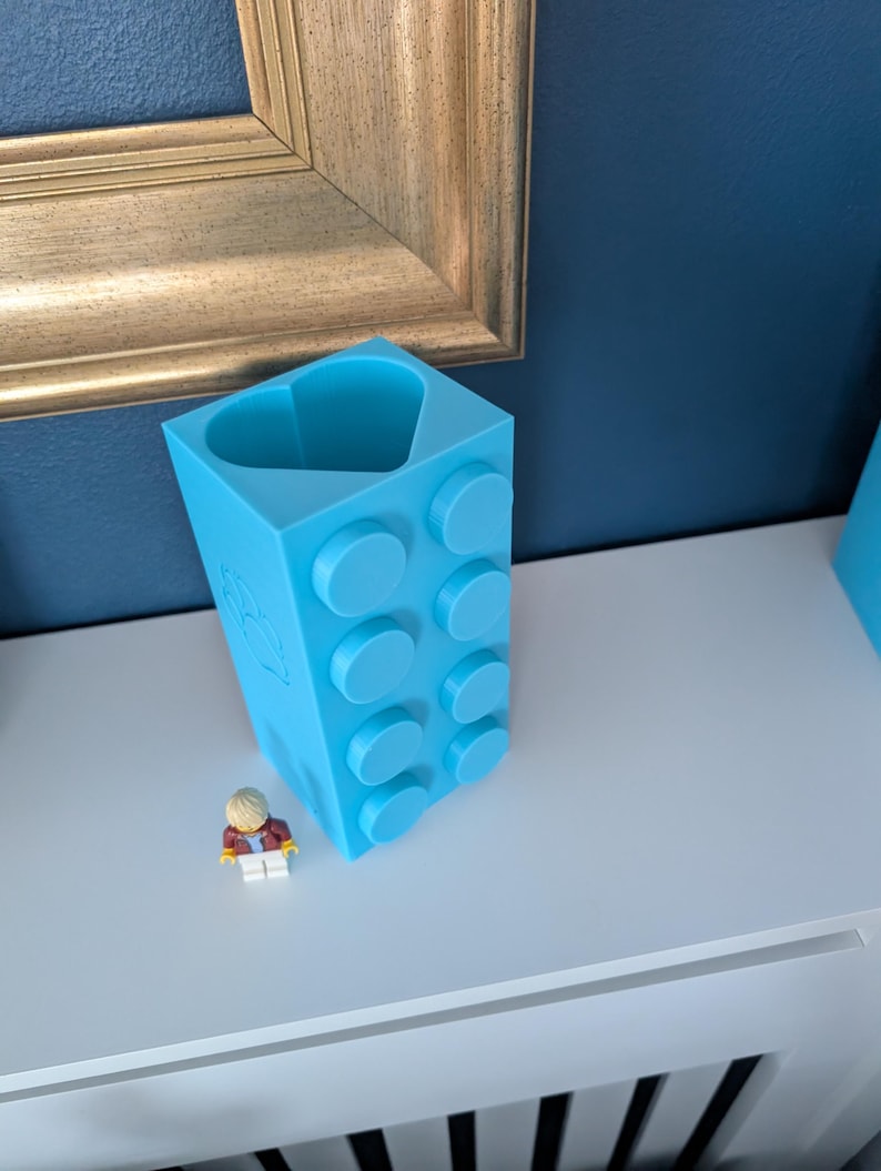 May include: A light blue, 3D-printed block-shaped container with circular studs, resembling a large building brick. A small Lego figure stands nearby. The container has a heart-shaped opening at the top.