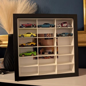 May include: A black framed display case with multiple compartments, showcasing a collection of toy cars. The cars are various colors, including red, blue, yellow, and green. The case is on a white surface against a dark blue wall.