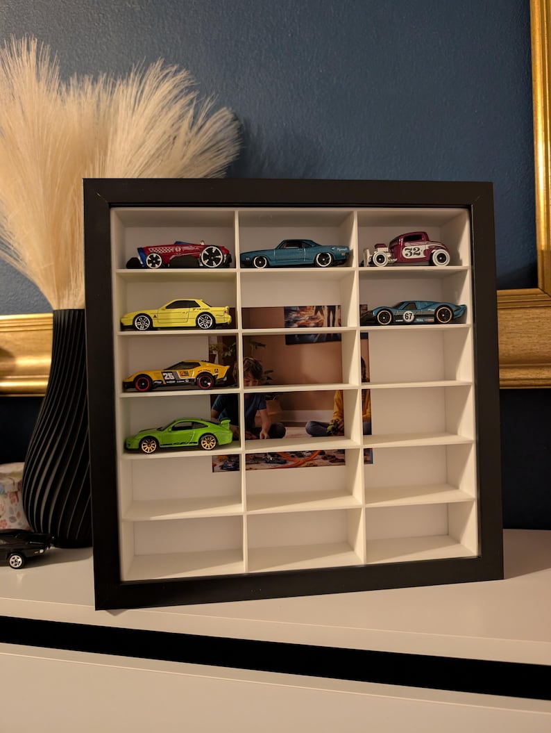 May include: A black framed display case with multiple compartments, showcasing a collection of toy cars. The cars are various colors, including red, blue, yellow, and green. The case is on a white surface against a dark blue wall.