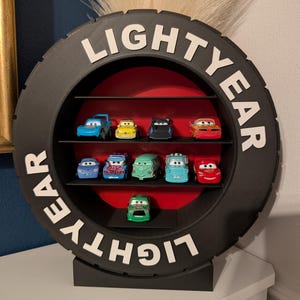 May include: A black tire-shaped shelf unit with the word "LIGHTYEAR" in white lettering. The shelf contains several toy cars of various colors, arranged on black shelves against a red backdrop. The unit is designed to stand on a flat surface.