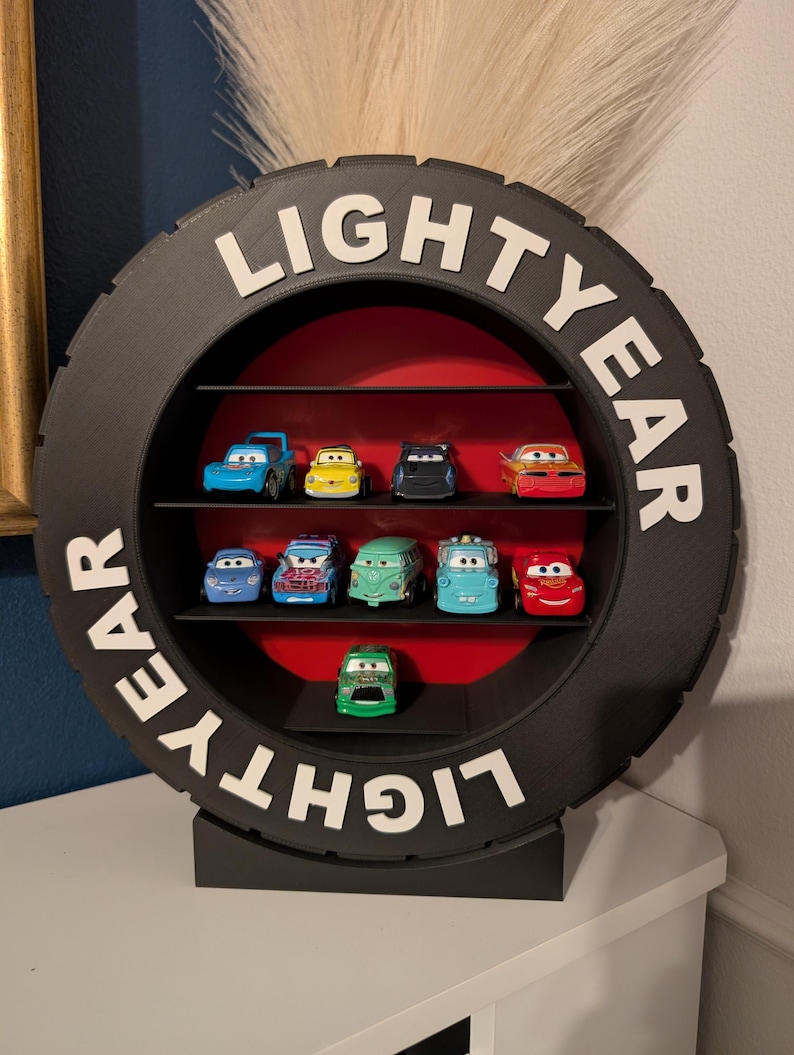 May include: A black tire-shaped shelf unit with the word "LIGHTYEAR" in white lettering. The shelf contains several toy cars of various colors, arranged on black shelves against a red backdrop. The unit is designed to stand on a flat surface.
