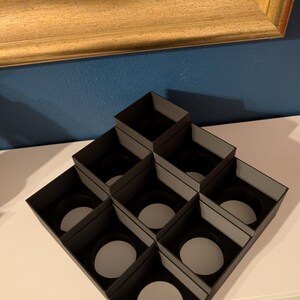 May include: A black, geometric display stand with nine square compartments, each holding a white circular object. The stand is on a white surface, with a gold-framed picture in the background.
