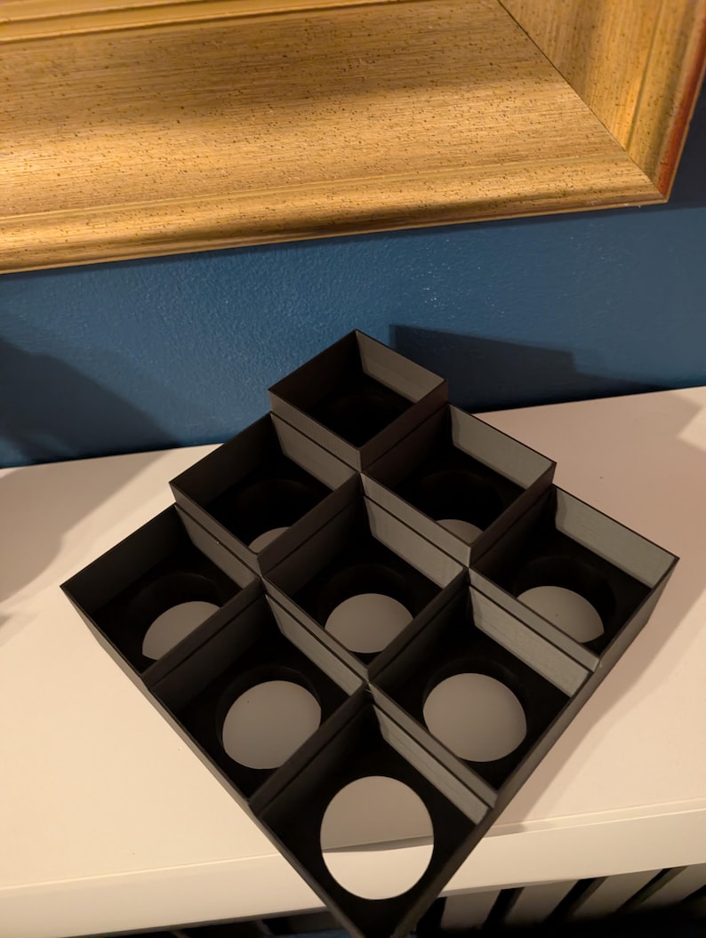 May include: A black, geometric display stand with nine square compartments, each holding a white circular object. The stand is on a white surface, with a gold-framed picture in the background.