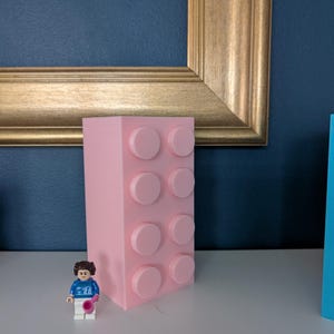 May include: A large, light pink building block with eight raised circular studs. A small Lego figure in a blue shirt and white pants stands in front of the block. The block is on a white surface with a gold framed picture in the background.