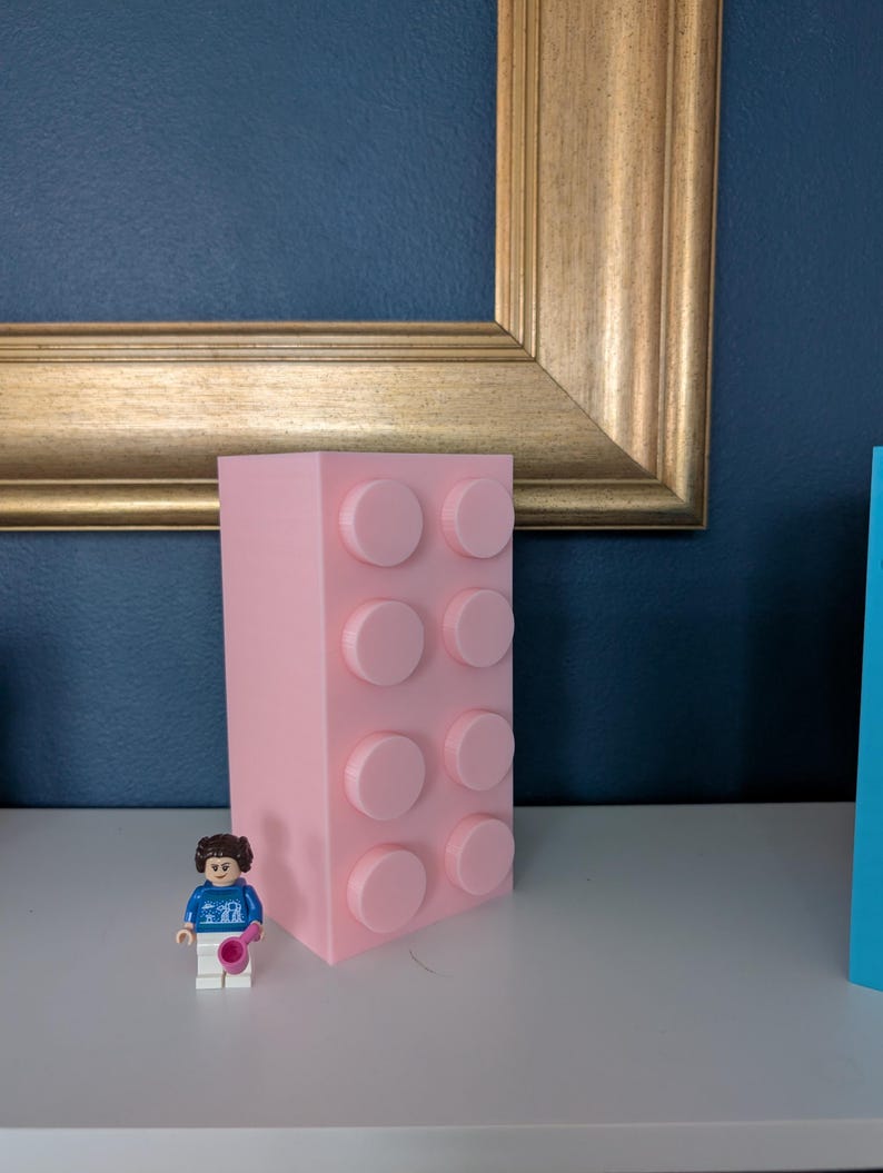 May include: A large, light pink building block with eight raised circular studs. A small Lego figure in a blue shirt and white pants stands in front of the block. The block is on a white surface with a gold framed picture in the background.