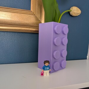 May include: A lavender-colored vase designed to resemble a building block, with a small Lego figure standing in front of it. The vase holds tulips with green stems and peach-colored blooms. The background is a dark blue wall.