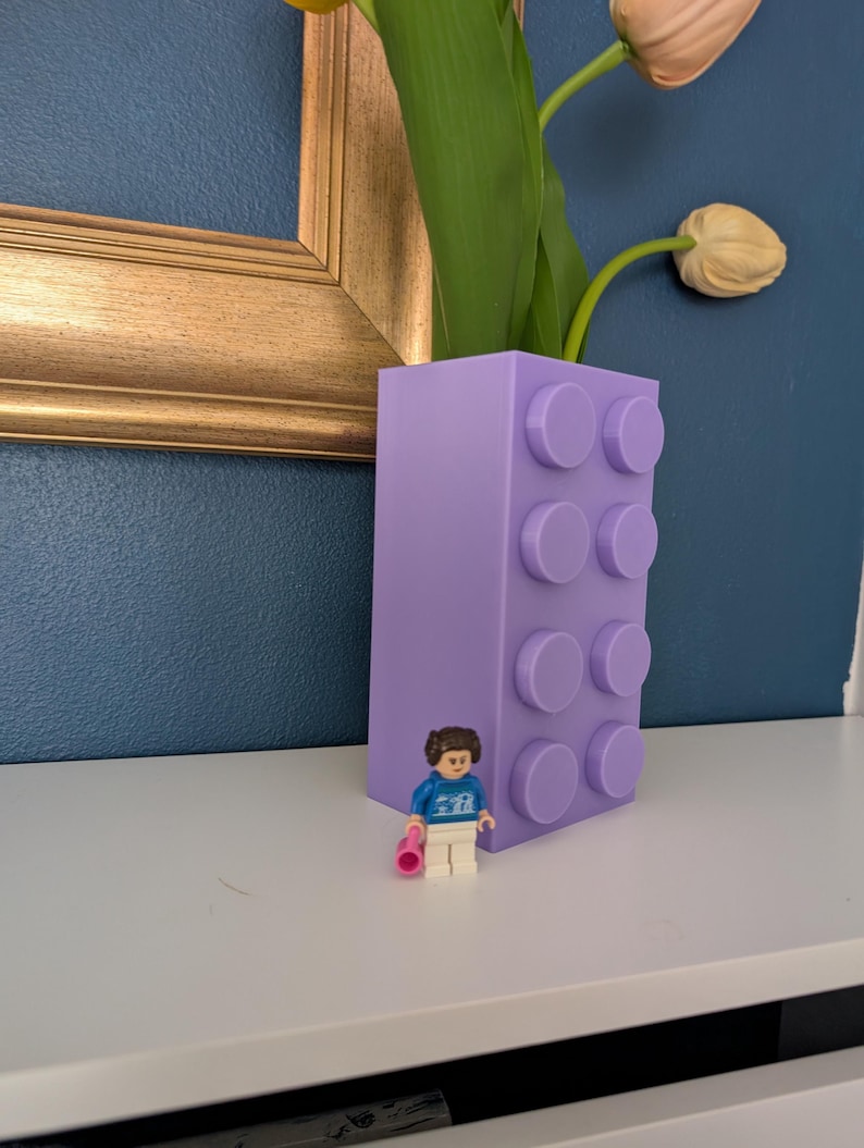 May include: A lavender-colored vase designed to resemble a building block, with a small Lego figure standing in front of it. The vase holds tulips with green stems and peach-colored blooms. The background is a dark blue wall.