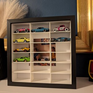 May include: A black framed display case with multiple compartments, showcasing a collection of toy cars. The cars are various colors, including red, blue, yellow, and green. The case is mounted on a white surface against a dark blue wall.