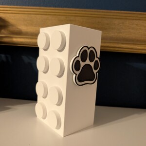May include: A white rectangular block with circular accents and a black and white paw print. The block is a decorative item, possibly a container or storage unit, with a playful design.