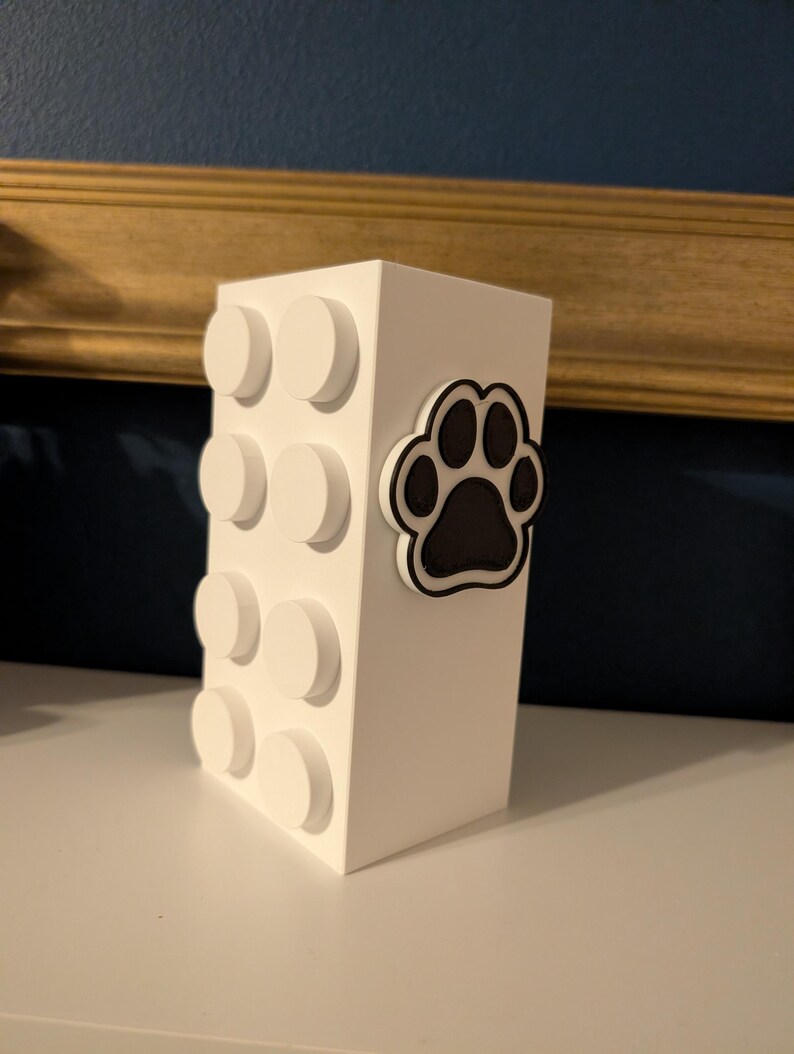 May include: A white rectangular block with circular accents and a black and white paw print. The block is a decorative item, possibly a container or storage unit, with a playful design.