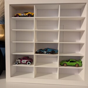 May include: A white display case with multiple compartments, showcasing toy cars in various colors. The cars include a yellow race car, a blue sports car, a maroon vintage car, and a green sports car. The case is designed for displaying collectibles.