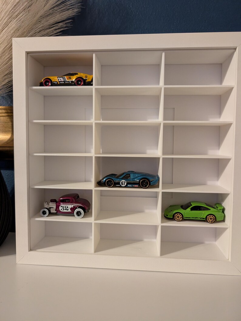 May include: A white display case with multiple compartments, showcasing toy cars in various colors. The cars include a yellow race car, a blue sports car, a maroon vintage car, and a green sports car. The case is designed for displaying collectibles.