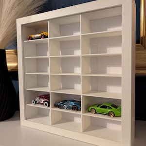 May include: A white, rectangular display case with multiple compartments, showcasing miniature toy cars. The case holds a yellow, a purple, a blue, and a green car. The cars are in various styles and colors, adding a playful touch to the display.