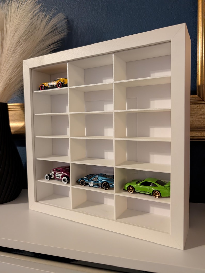 May include: A white, rectangular display case with multiple compartments, showcasing miniature toy cars. The case holds a yellow, a purple, a blue, and a green car. The cars are in various styles and colors, adding a playful touch to the display.