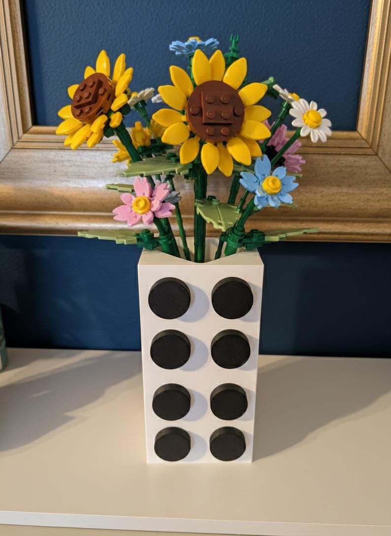 May include: A white rectangular vase with eight black circular accents holds a bouquet of colorful flowers. The flowers include yellow sunflowers, pink, blue, and white blossoms, all constructed from interlocking plastic bricks.