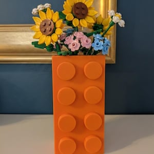 May include: An orange brick-shaped vase filled with colorful Lego flowers. The flowers include yellow sunflowers with brown centers, pink and blue flowers, and white accents. The vase has six orange studs.