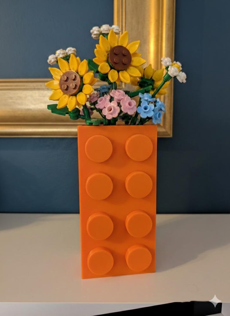 May include: An orange brick-shaped vase filled with colorful Lego flowers. The flowers include yellow sunflowers with brown centers, pink and blue flowers, and white accents. The vase has six orange studs.