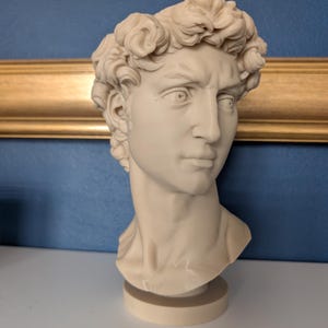 May include: A cream-colored bust of a man with curly hair, a stern expression, and a detailed face. The sculpture is on a round base, set against a blue wall with a gold-colored frame.