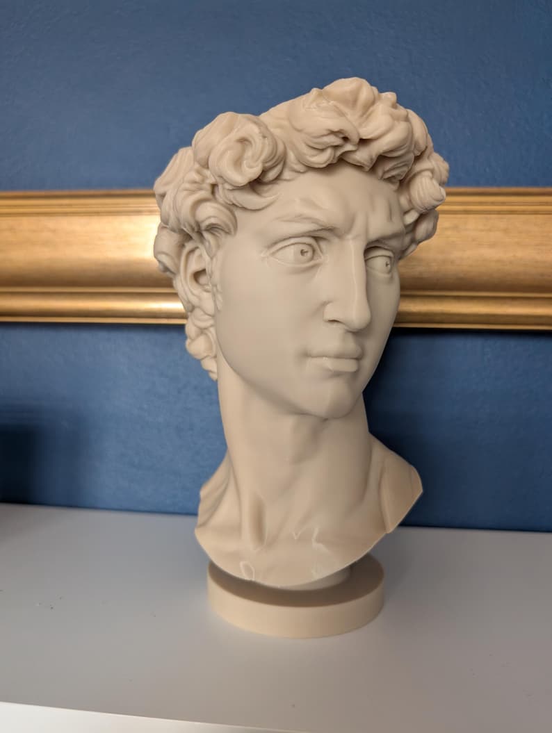 May include: A cream-colored bust of a man with curly hair, a stern expression, and a detailed face. The sculpture is on a round base, set against a blue wall with a gold-colored frame.