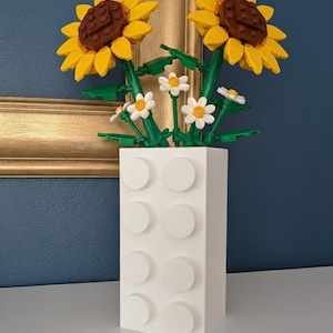 May include: A white vase made of interlocking blocks holds a bouquet of sunflowers and daisies. The sunflowers have yellow petals and brown centres. The daisies are white with yellow centres. The vase is on a white surface with a gold-framed picture in the background.