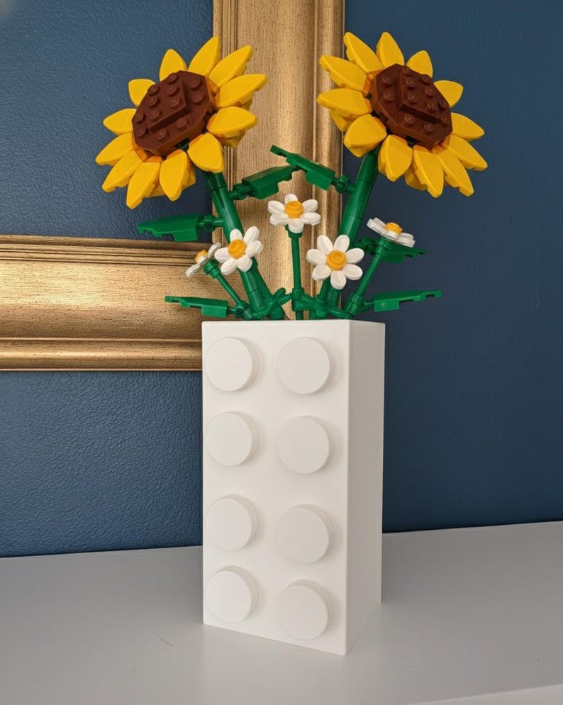 May include: A white vase made of interlocking blocks holds a bouquet of sunflowers and daisies. The sunflowers have yellow petals and brown centers. The daisies are white with yellow centers. The vase is on a white surface with a gold-framed picture in the background.