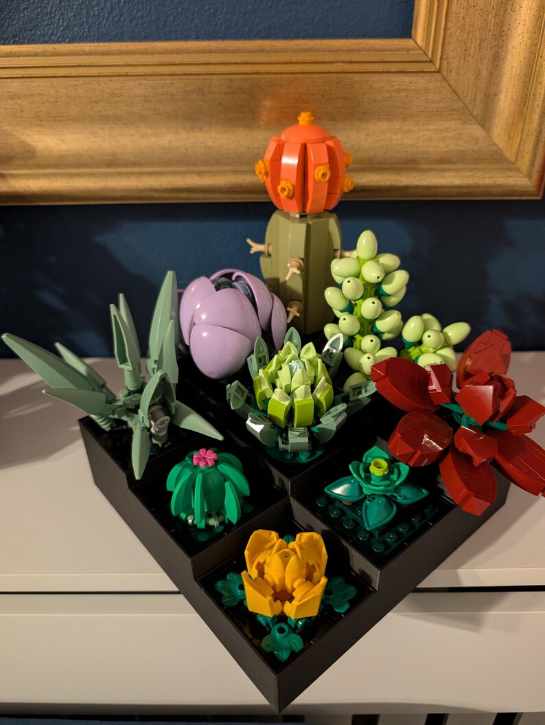 May include: A collection of colorful Lego succulent plants in black square planters. The plants include a green agave, a purple flower, a green cactus with a pink flower, a yellow flower, and a red flower. The background includes a gold-framed picture and a blue wall.