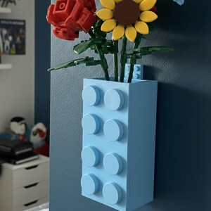 Op de afbeelding: Een lichtblauwe vaas gemaakt van speelgoedbouwstenen, met kleurrijke speelgoedbloemen. De bloemen zijn onder meer een rode roos, een gele zonnebloem en kleine blauwe bloemen. De vaas is bevestigd aan een donkerblauwe muur.