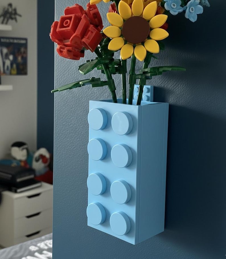 Op de afbeelding: Een lichtblauwe vaas gemaakt van speelgoedbouwstenen, met kleurrijke speelgoedbloemen. De bloemen zijn onder meer een rode roos, een gele zonnebloem en kleine blauwe bloemen. De vaas is bevestigd aan een donkerblauwe muur.