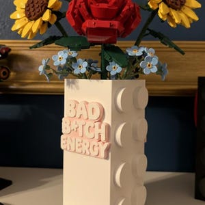 May include: A white vase with a Lego-like design, holding a bouquet of Lego flowers. The bouquet includes yellow sunflowers, a red rose, and blue forget-me-nots. The vase has the text "BAD B*TCH ENERGY" in pink.