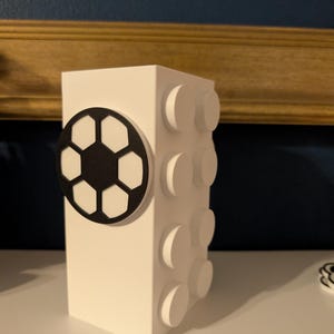 May include: A white, rectangular block-shaped decorative item with a black and white soccer ball design on one side and raised circular details on the other. The item is on a white surface.