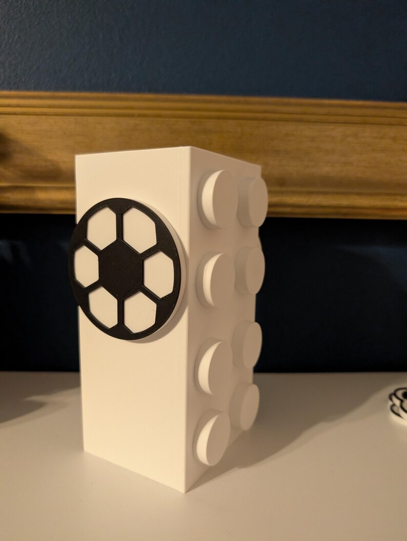 May include: A white, rectangular block-shaped decorative item with a black and white soccer ball design on one side and raised circular details on the other. The item is on a white surface.