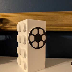 May include: A white rectangular block with circular white studs on one side and a black and white soccer ball design on the front. The block is a decorative item, possibly a storage container or a decorative piece.