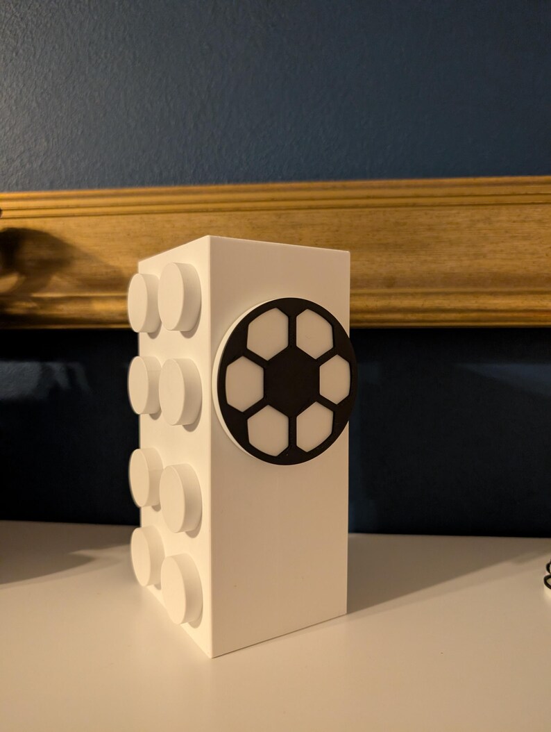 May include: A white rectangular block with circular white studs on one side and a black and white soccer ball design on the front. The block is a decorative item, possibly a storage container or a decorative piece.
