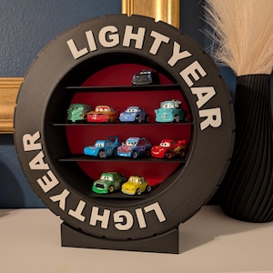 May include: A black, tire-shaped shelf with the word "LIGHTYEAR" in white letters. The shelf displays toy cars of various colors. The interior of the shelf is red. The shelf is on a white surface.
