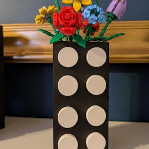 Puede incluir: Un jarr&oacute;n rectangular negro con detalles circulares blancos, que contiene un ramo de flores coloridas. Las flores incluyen rosas rojas, girasoles amarillos, nomeolvides azules y tulipanes morados. El jarr&oacute;n es de pl&aacute;stico.