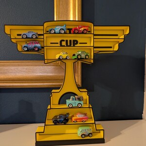 Puede incluir: Una estanter&iacute;a amarilla con forma de trofeo y ribete negro, que exhibe coches de juguete en miniatura. La palabra "CUP" est&aacute; impresa en la estanter&iacute;a. La estanter&iacute;a est&aacute; contra una pared azul oscuro y un cuadro con marco dorado.