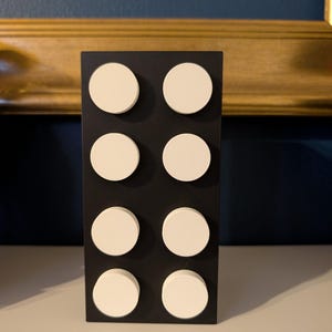 Puede incluir: Un objeto decorativo rectangular negro con ocho acentos circulares blancos dispuestos en dos columnas de cuatro. El objeto est&aacute; de pie sobre una superficie blanca, con una pared azul oscuro y una obra de arte con marco dorado en el fondo.