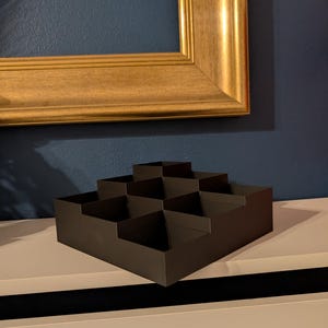 May include: A dark brown, stepped display box with multiple compartments. The box is made of a matte material and sits on a white surface. A gold-colored picture frame hangs in the background.