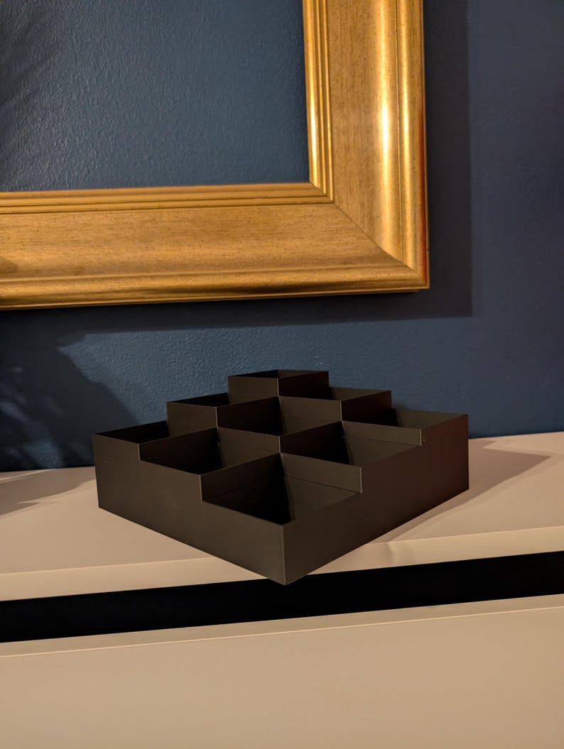 May include: A dark brown, stepped display box with multiple compartments. The box is made of a matte material and sits on a white surface. A gold-colored picture frame hangs in the background.