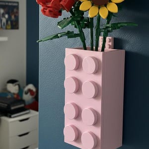 Op de afbeelding: Een lichtroze vaas gemaakt van bouwstenen, met kleurrijke kunstbloemen. De bloemen omvatten een rode roos, een gele zonnebloem en blauwe bloemen. De vaas is bevestigd aan een blauwe muur.