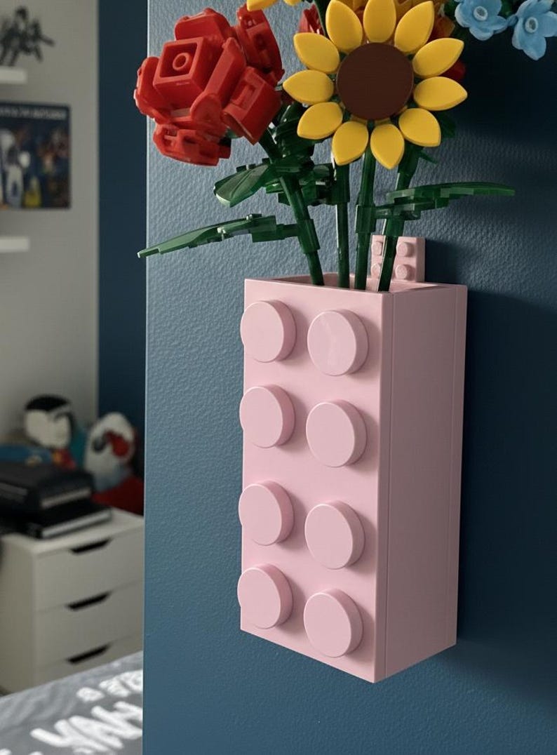 Op de afbeelding: Een lichtroze vaas gemaakt van bouwstenen, met kleurrijke kunstbloemen. De bloemen omvatten een rode roos, een gele zonnebloem en blauwe bloemen. De vaas is bevestigd aan een blauwe muur.